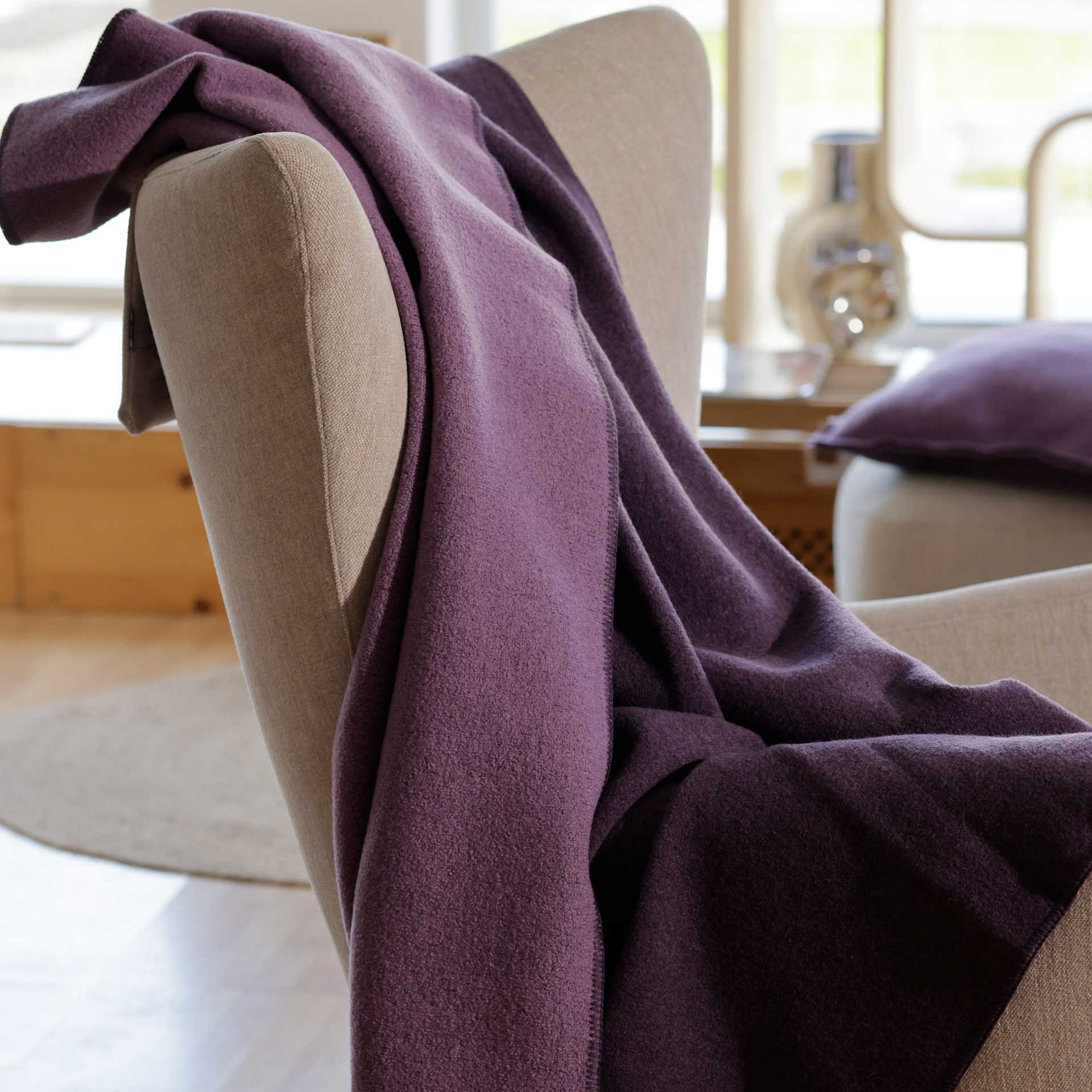 disana Doubleface-Decke in Aubergine-Flieder aus Bio-Merinowolle auf einem beigen Sessel in einem hellen Wohnzimmer